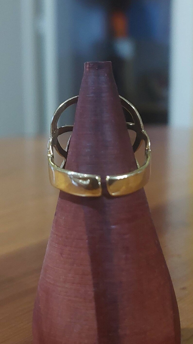 Puede incluir: Un anillo de tono dorado con un dise&ntilde;o geom&eacute;trico. El anillo se muestra en un soporte de anillo de madera.