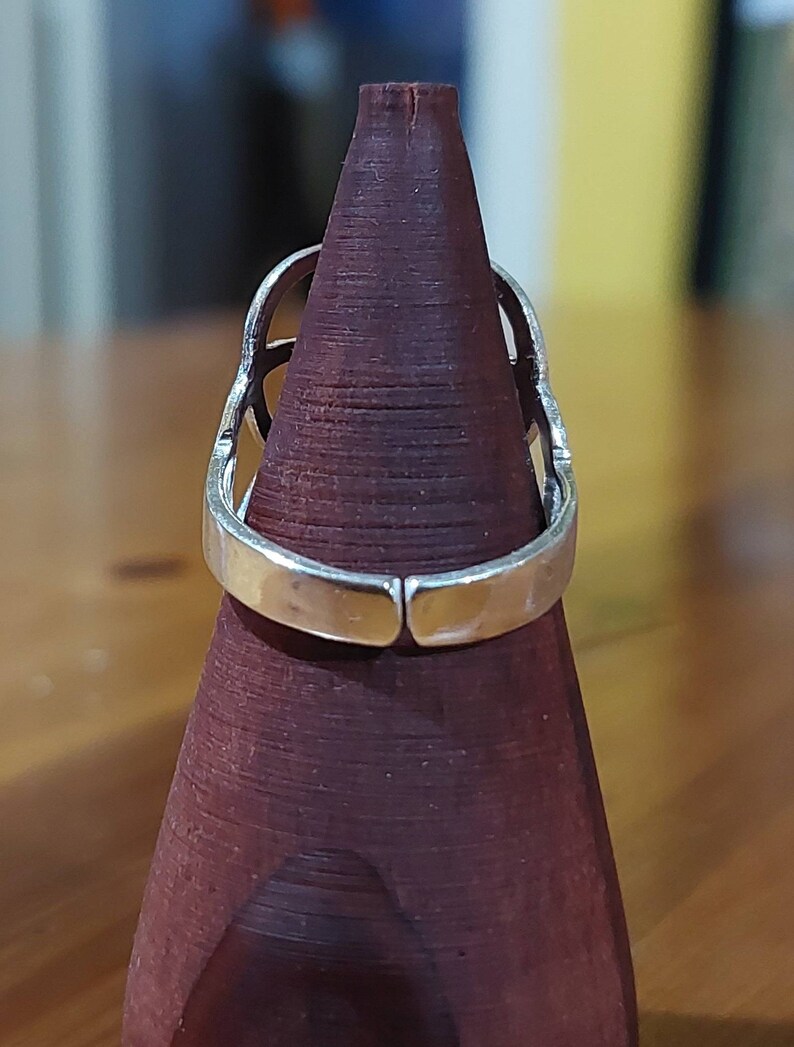 Puede incluir: Un anillo de plata con un dise&ntilde;o geom&eacute;trico. El anillo se muestra en un soporte de anillo de madera.