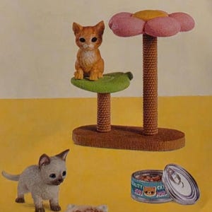 May include: A toy set labeled KITTENS NO.1 featuring an orange kitten figurine on a green leaf platform with a pink flower top, a grey kitten figurine, a small white cat-shaped food bowl, and an open metal can of cat food with a blue label.