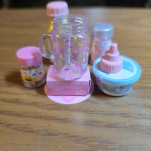 Puede incluir: Una colección de alimentos de juguete en miniatura, que incluye un tarro transparente con chispas de colores, una pequeña caja rosa con el texto "Just Baked" y varios recipientes con tapas rosas. Los artículos están dispuestos sobre una superficie de madera.