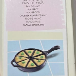 May include: A black cast iron skillet divided into eight triangular sections, each containing a piece of yellow cornbread topped with a small green garnish. Above the skillet, a light pink paper note lists the word cornbread in many languages.