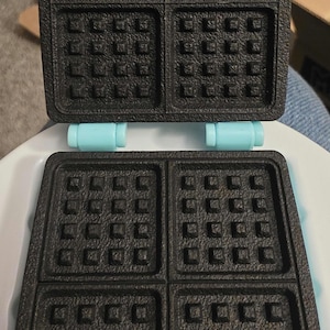 May include: A black waffle maker with a square design, open to reveal the cooking surfaces. The top and bottom sections are connected by light blue hinges. Each cooking surface is divided into four square sections.