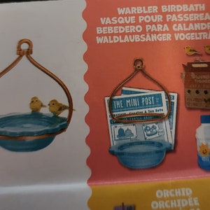 May include: A miniature birdbath with two yellow birds perched on the edge. The birdbath is teal with a copper-colored metal frame. The image also shows a newspaper, a small carton, and a bottle.