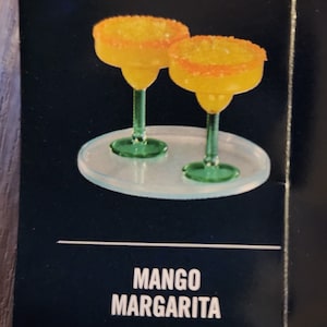 May include: Two yellow mango margaritas in clear glass margarita glasses with a sugared rim, sitting on a clear glass coaster. The text "MANGO MARGARITA" is printed below the glasses.