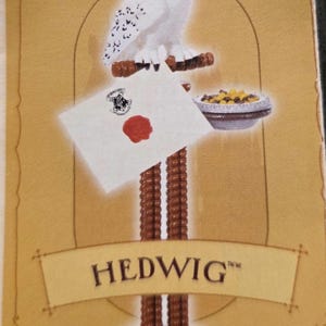 May include: A figurine of Hedwig, a snowy owl, perched on a brown branch. Below, a white envelope with a red wax seal and a Hogwarts crest rests on a stand. A small bowl of food is also present. The base is brown, and the background is tan.