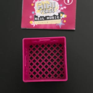 May include: A bright pink square crate with a lattice design sits below a pink card. The card reads "MINI VERSE REAL MUSIC" in white and yellow text, with a number one in a circle.