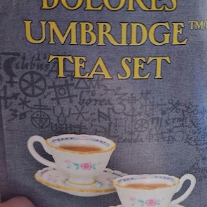 May include: A tea set featuring two teacups and saucers, filled with tea. The cups and saucers are white with pink floral designs and blue and gold trim. The text "DOLORES UMBRIDGE TEA SET" is displayed in gold lettering.