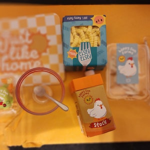 May include: A collection of miniature food items, including pasta, stock, and a bowl with a spoon. The pasta is in a blue box with the text "100% Happy Pasta". The stock box features a cartoon chicken. The items are arranged on a yellow surface.