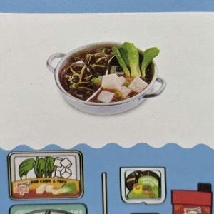 May include: A toy hot pot set featuring a silver dual-compartment pot filled with dark broth, tofu, and bok choy. Included are a ladle, a container labeled Bok Choy & Tofu, a container of chives and peppercorns, and a red carton labeled Spicy Broth.