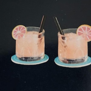 May include: Two identical short glass tumblers filled with a pale orange liquid, each containing a dark straw and garnished with a slice of pink grapefruit. Each glass rests on a small, round, light blue coaster against a solid black background.