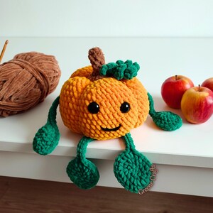 Op de afbeelding: Een handgemaakte oranje gehaakte pompoen met groene bladeren en benen, zwarte ogen en een lachend gezicht. Een bol bruin garen en drie rode appels staan ook op de foto. De pompoen is een herfstthema decoratie.