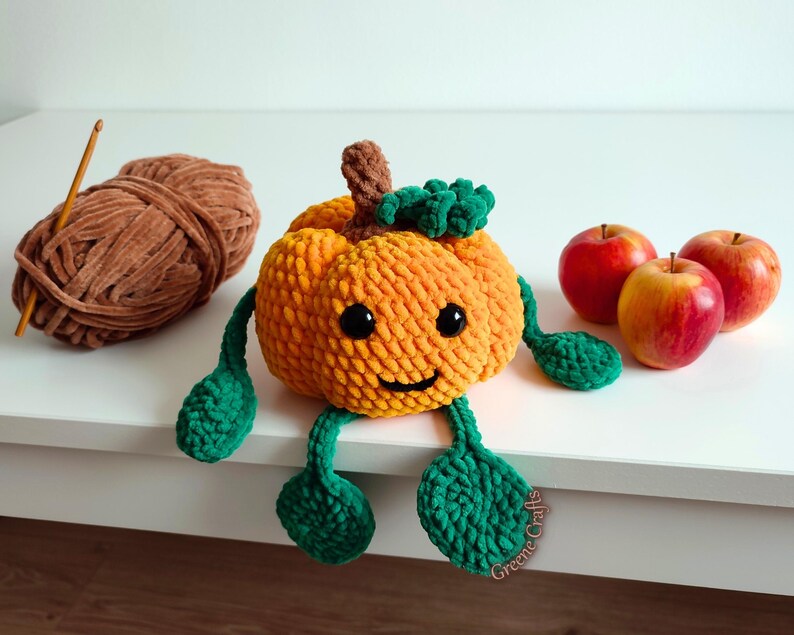 Op de afbeelding: Een handgemaakte oranje gehaakte pompoen met groene bladeren en benen, zwarte ogen en een lachend gezicht. Een bol bruin garen en drie rode appels staan ook op de foto. De pompoen is een herfstthema decoratie.