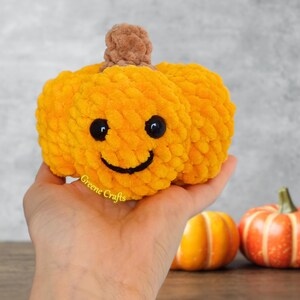 Op de afbeelding: Een geel-oranje, lachende pompoen van pluche met zwarte ogen en een gestikte mond. De pompoen heeft een bruine steel en wordt in een hand gehouden. Twee andere pompoenen staan op de achtergrond. De tekst "Greene Crafts" is zichtbaar.