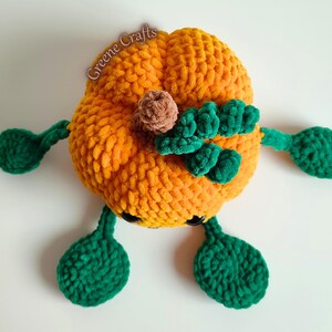 Op de afbeelding: Een gehaakte oranje pompoen met groene bladeren, armen en voeten. Het heeft een bruine stengel en zwarte ogen. De tekst "Greene Crafts" is zichtbaar. Perfect voor herfstdecoratie en seizoensgebonden displays.