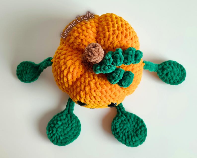 Op de afbeelding: Een gehaakte oranje pompoen met groene bladeren, armen en voeten. Het heeft een bruine stengel en zwarte ogen. De tekst "Greene Crafts" is zichtbaar. Perfect voor herfstdecoratie en seizoensgebonden displays.