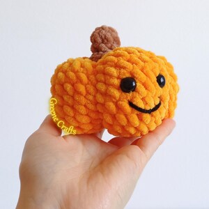 Op de afbeelding: Een handgemaakte oranje gehaakte pompoen met een lachend gezicht en een bruine stengel. De pompoen wordt in een hand gehouden tegen een witte achtergrond. De pompoen is een knuffel.
