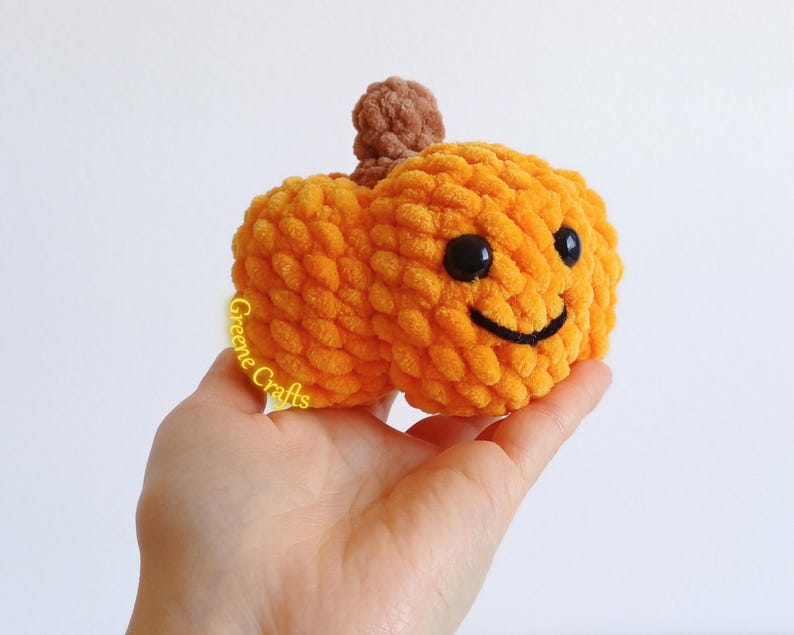 Op de afbeelding: Een handgemaakte oranje gehaakte pompoen met een lachend gezicht en een bruine stengel. De pompoen wordt in een hand gehouden tegen een witte achtergrond. De pompoen is een knuffel.