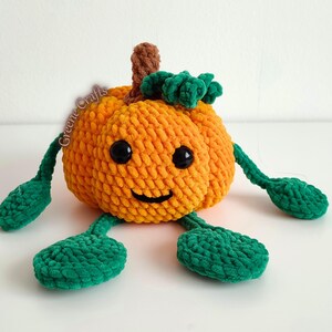 Op de afbeelding: Een gehaakte oranje pompoen met een lachend gezicht, zwarte ogen en groene bladeren en stelen. De pompoen heeft groene armen en voeten. De tekst "Greene Crafts" is zichtbaar op de zijkant van de pompoen.