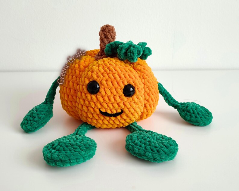 Op de afbeelding: Een gehaakte oranje pompoen met een lachend gezicht, zwarte ogen en groene bladeren en stelen. De pompoen heeft groene armen en voeten. De tekst "Greene Crafts" is zichtbaar op de zijkant van de pompoen.