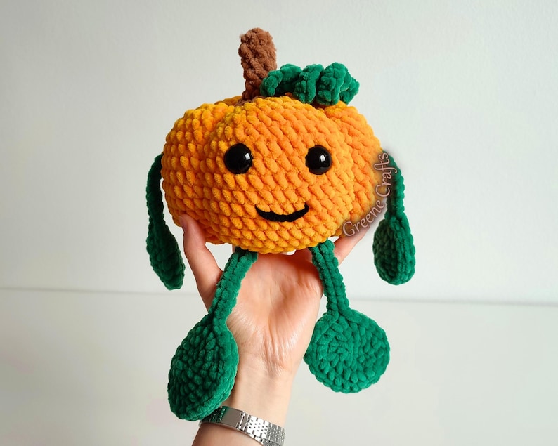 Op de afbeelding: Een handgemaakt pompoenspeeltje met een lachend gezicht, zwarte ogen en groene bladvormige benen. De pompoen is oranje met een bruine stengel en groene bladeren bovenop. Het speelgoed wordt in een hand gehouden.