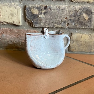 May include: A white ceramic pitcher with a speckled glaze and a handle. The pitcher has a rounded shape and a small spout.
