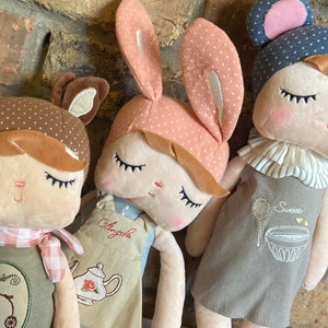 May include: Three plush dolls with embroidered details. The doll on the left is a brown deer with a pink and white checked scarf and a green dress with a bicycle design. The middle doll is a pink bunny with a blue and white apron and the text "Angel" embroidered on it. The doll on the right is a grey mouse with a blue hat and a grey dress with the text "Sweet" embroidered on it.