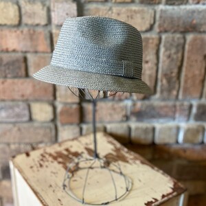 Pode incluir: Um chapéu fedora cinza tecido em um suporte de exibição de metal. O chapéu tem uma aba larga e uma coroa ligeiramente afundada.