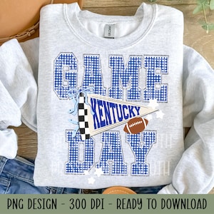 May include: A light grey crewneck sweatshirt featuring the text GAME DAY in a blue and white gingham pattern. A blue and white pennant with the word KENTUCKY is centred, accompanied by a brown football graphic and a small blue bow accent.