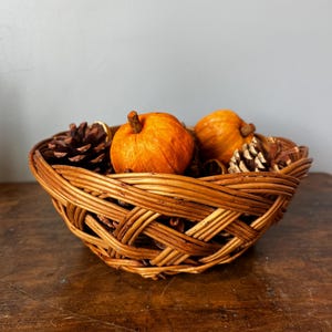 May include: A round, woven brown wicker basket filled with two small orange pumpkins and two brown pinecones. The basket features a crisscross pattern of thin, flexible reeds, creating a sturdy, open-weave structure that holds the seasonal items.