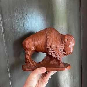 May include: A reddish-brown carved figurine of a bison standing on a flat base. The sculpture features detailed textures representing the animal's thick fur on the head and shoulders, with smooth, carved surfaces along the back and hindquarters.
