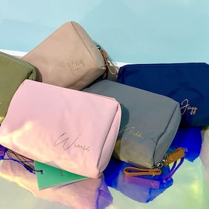 May include: Five different coloured zippered pouches with gold lettering. The pouches are in shades of green, beige, pink, blue, and grey. The lettering on the pouches reads "Jenny", "Kay", "Winnie", "Adele", and "Jazz".
