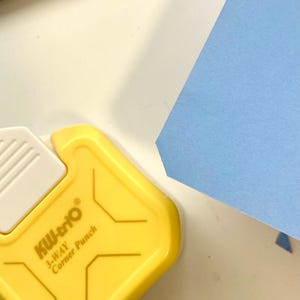 May include: A yellow KW-trio 3-Way Corner Punch tool with white accents and the text "3-WAY Corner Punch". A light blue piece of paper is in the background.