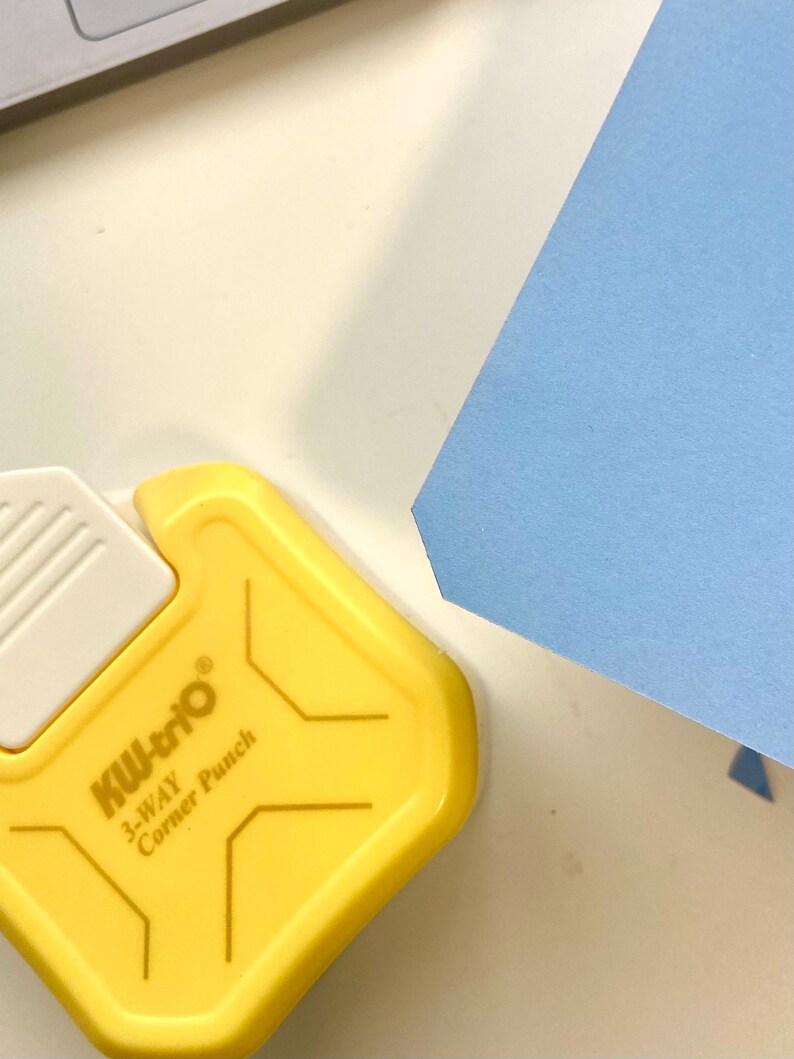 May include: A yellow KW-trio 3-Way Corner Punch tool with white accents and the text "3-WAY Corner Punch". A light blue piece of paper is in the background.