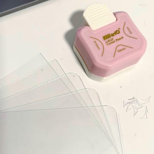 May include: A pink and white corner punch with the text "Kili-to 3-WAY Corner Punch" and clear plastic sheets. The punch has measurements of R10mm and R6mm. Small cut pieces of plastic are visible.