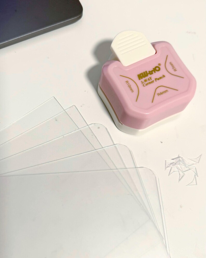 May include: A pink and white corner punch with the text "Kili-to 3-WAY Corner Punch" and clear plastic sheets. The punch has measurements of R10mm and R6mm. Small cut pieces of plastic are visible.