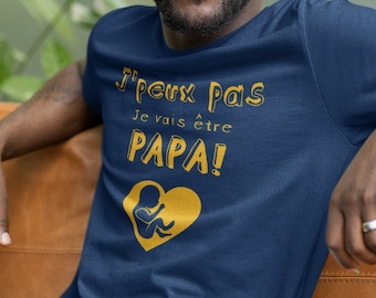 May include: A man wearing a navy blue t-shirt with the text "J'peux pas je vais être PAPA!" in gold lettering and a gold graphic of a baby in a heart.