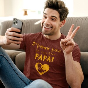 May include: A man wearing a maroon t-shirt with the text "J'Peux Pas Je Vais &Ecirc;tre Papa!" in yellow. The shirt also features a yellow graphic of a heart with a baby inside. He is sitting on the floor and holding a black smartphone.