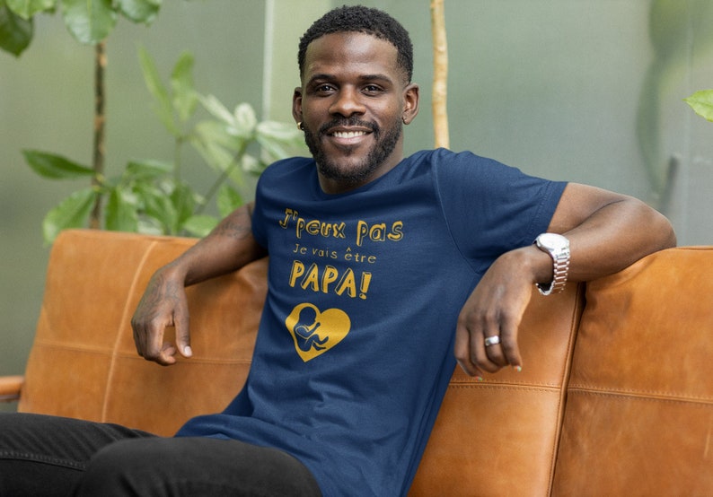 May include: A man wearing a navy blue t-shirt with the text "J'peux pas je vais &ecirc;tre PAPA!" in gold lettering and a gold graphic of a baby in a heart.