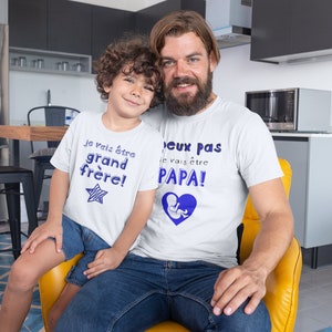 May include: Two people wearing white t-shirts with blue text. The text on the smaller shirt says "Je vais &ecirc;tre grand fr&egrave;re!" and the text on the larger shirt says "J'eux pas je vais &ecirc;tre PAPA!"