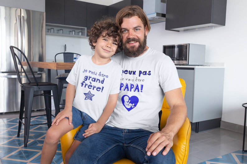 May include: Two people wearing white t-shirts with blue text. The text on the smaller shirt says "Je vais &ecirc;tre grand fr&egrave;re!" and the text on the larger shirt says "J'eux pas je vais &ecirc;tre PAPA!"