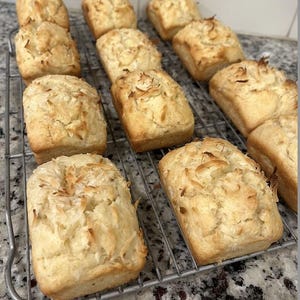 May include: A close-up of a wire rack with 12 small, golden brown loaves of bread topped with shredded coconut. The loaves are arranged in rows on the rack.