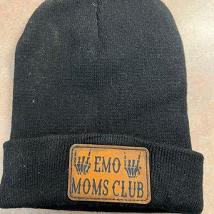 Op de afbeelding: Zwarte gebreide beanie met een bruine leren patch waarop "EMO MOMS CLUB" staat met twee skelethandjes die de rock-and-roll handbeweging maken.