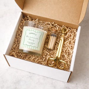 May include: A gift box containing a candle, matches, and wick trimmer. The candle has a label that reads "EMMA'S Bachelorette Party" with a light green and white striped design. The matches are in a small glass bottle with a cork. The wick trimmer is gold.