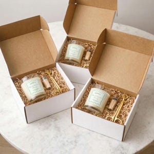 May include: Three gift boxes, each containing a candle in a glass jar, matches, and gold-coloured scissors. The candle jars have labels reading "Emma's Bachelorette Party". The boxes are filled with shredded paper.