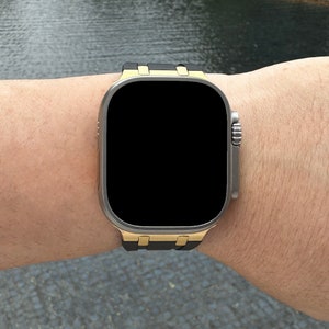 Może przedstawiać: Czarny i złoty smartwatch z czarnym paskiem na nadgarstku osoby.
