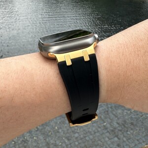 Może przedstawiać: Srebrny zegarek Apple Watch z czarnym paskiem z gumy i złotymi akcentami. Zegarek jest na nadgarstku osoby.