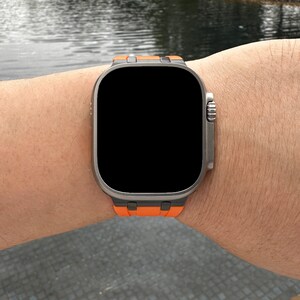 Puede incluir: Un reloj inteligente negro con marco plateado y una banda de silicona naranja. La esfera del reloj es negra y est&aacute; en blanco.