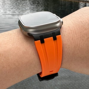Puede incluir: Un reloj inteligente plateado con una banda de silicona negra y naranja. La esfera del reloj es negra y la banda tiene un dise&ntilde;o texturizado.