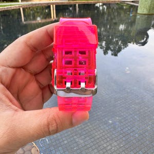 May include: A bright pink watch band with a silver buckle. The band is made of a translucent material and has a grid pattern.