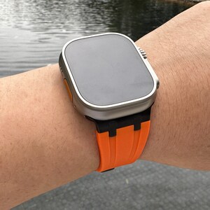 Puede incluir: Un reloj inteligente plateado con una banda de silicona negra y naranja. La esfera del reloj es negra y rectangular.